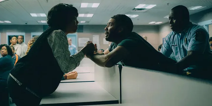 Two people grip hands intensely across a counter in what appears to be a prison visitation room.