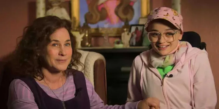 Two women sit together in a decorated room, one wearing a pink cancer patient cap and glasses.