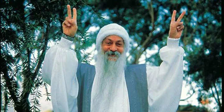 An elderly bearded man in white robes raises both hands in peace signs outdoors among trees.