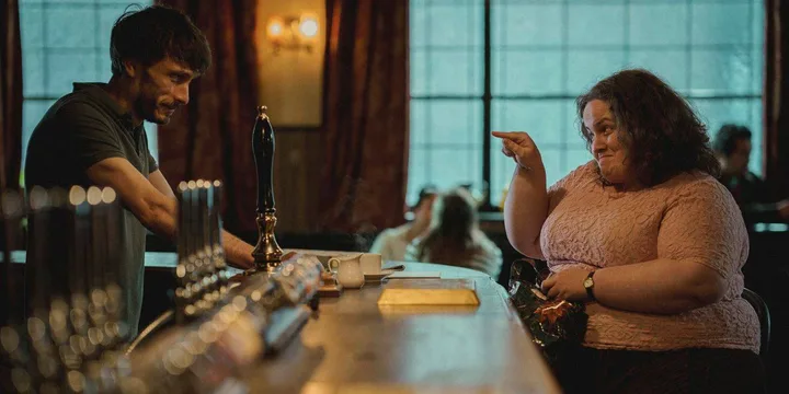 A man and woman have an animated conversation at a British pub bar, warm atmospheric lighting.