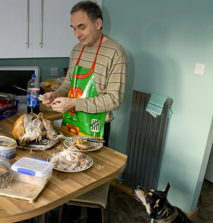 Middle-aged man in apron carving a roasted bird in a home kitchen while a dog watches eagerly.