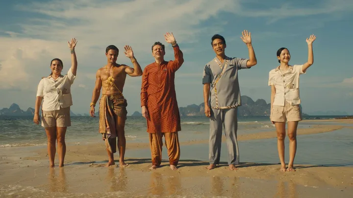 Five cast members in colorful costumes wave on a tropical beach with dramatic cliffs behind them.