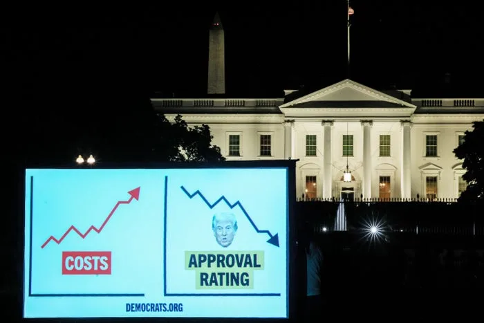 Nighttime billboard in front of the White House showing rising costs and Trump's falling approval rating chart.