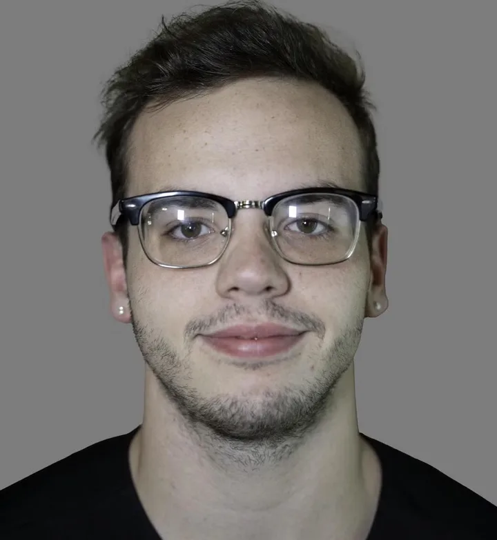 Headshot portrait of a young man with glasses and stubble against a plain gray background.