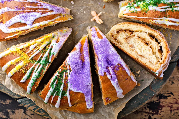 Sliced king cake with purple, gold, and green sugar decorations and white icing, a plastic baby visible nearby.