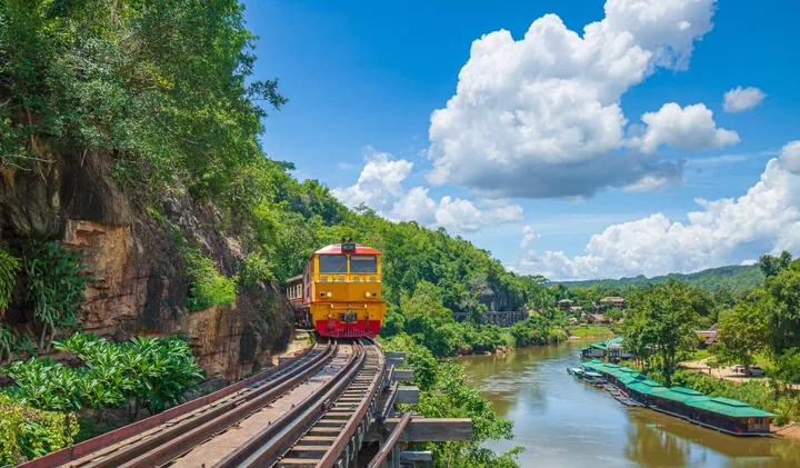 Yellow train on a scenic railway hugging a cliff beside a river in a lush Thai jungle.