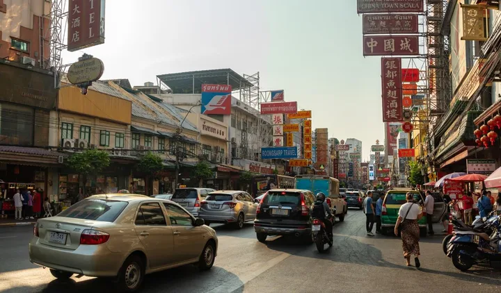 Busy Bangkok Chinatown street with heavy traffic, motorcycles, and colorful commercial signage.