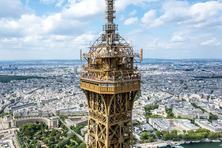 Aerial close-up of the Eiffel Tower's upper levels with Paris cityscape stretching into the distance.