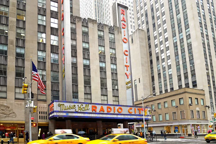 Exterior street-level view of Radio City Music Hall in New York City with yellow taxis passing by.