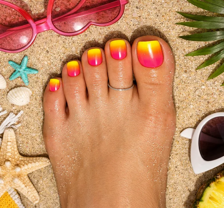 Close-up of a foot with sunset ombre pedicure (yellow to pink) on sandy beach with starfish and sunglasses.