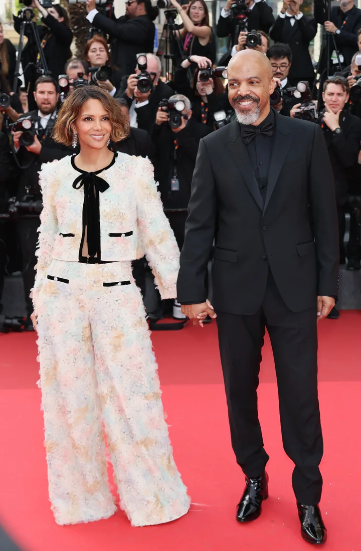 A couple holding hands on a red carpet, woman in floral suit, man in black tuxedo, paparazzi behind them.