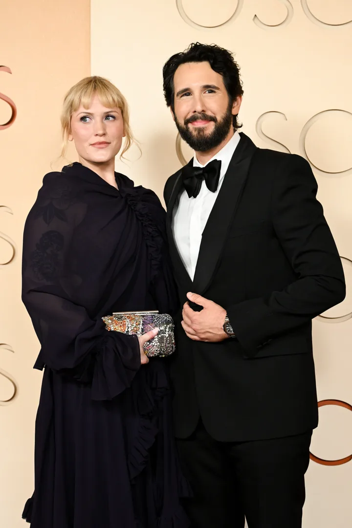 A couple in formal black attire posing together at an Oscars event, smiling warmly.