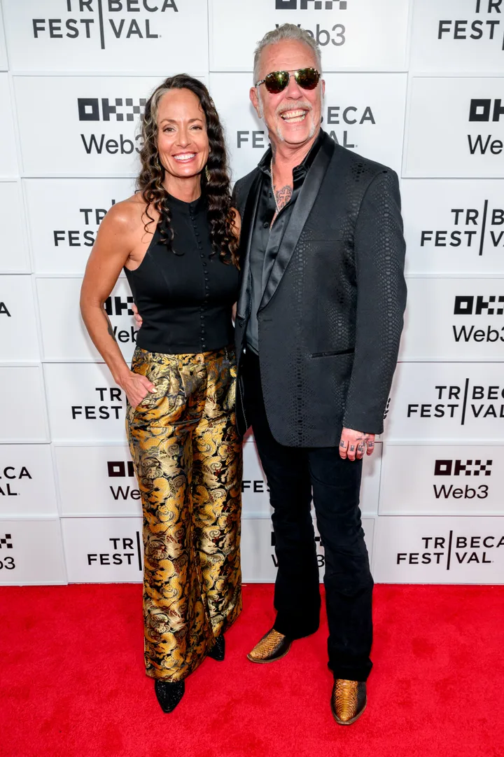 A couple smiling together on a Tribeca Festival red carpet, woman in gold pants, man in black jacket.