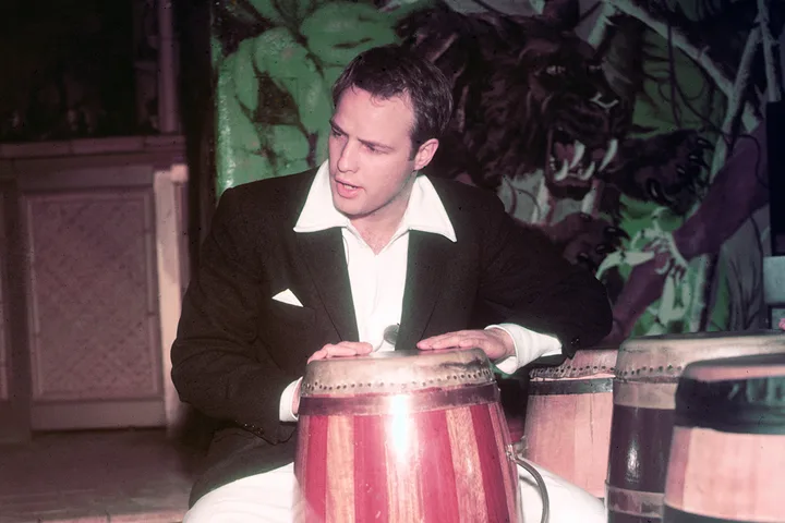 Young man in a black suit playing conga drums before a painted jungle mural backdrop.