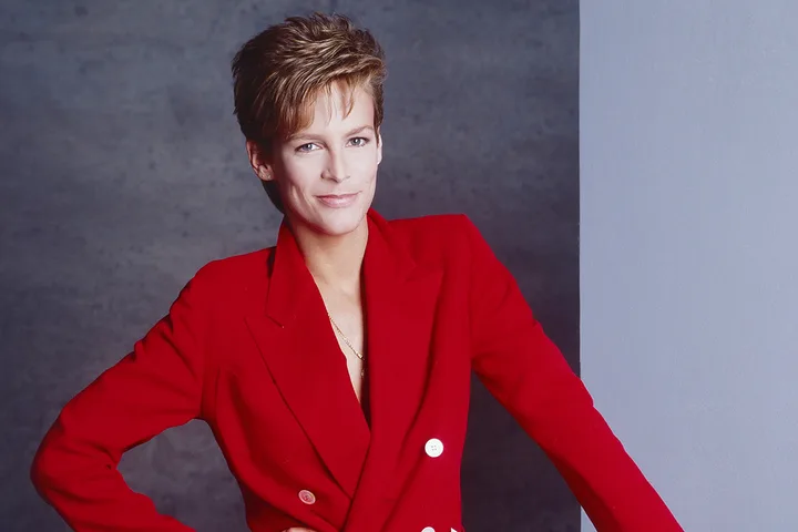Woman with short hair in a red blazer poses confidently against a grey studio background.