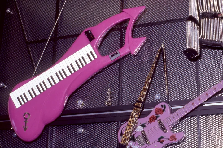 Bright pink keytar with Prince's love symbol displayed alongside a pink guitar on a dark wall.