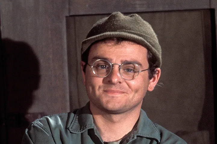 Man in military cap and round glasses smiling warmly in a dimly lit indoor setting.