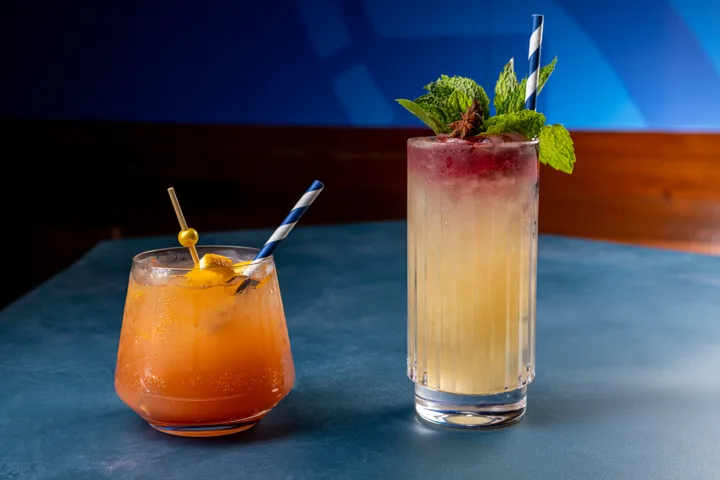 Two cocktails with garnishes on a blue bar surface in dim lounge lighting.