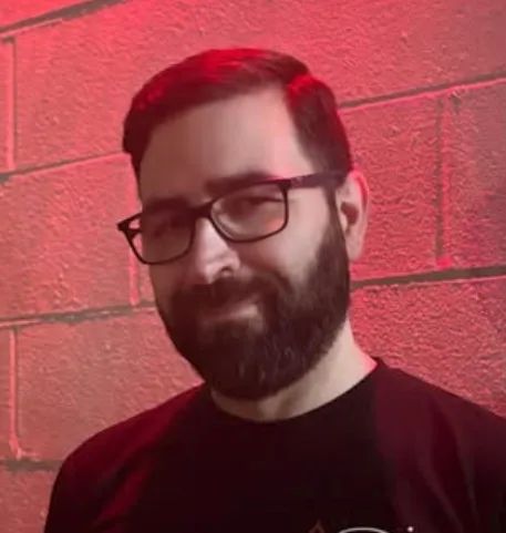 Bearded man with glasses smiling against a red brick wall in dim lighting.