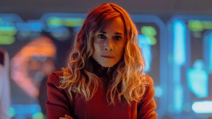 Woman in red Star Trek uniform on a sci-fi bridge set, arms crossed, serious expression.