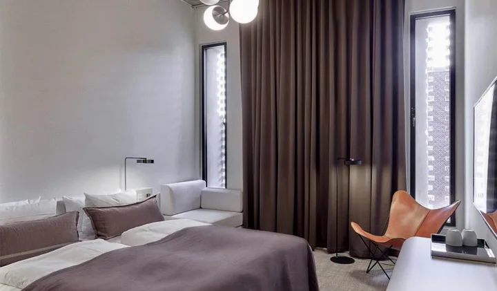 Modern hotel bedroom with dark curtains, globe pendant light, leather butterfly chair, and neutral tones.
