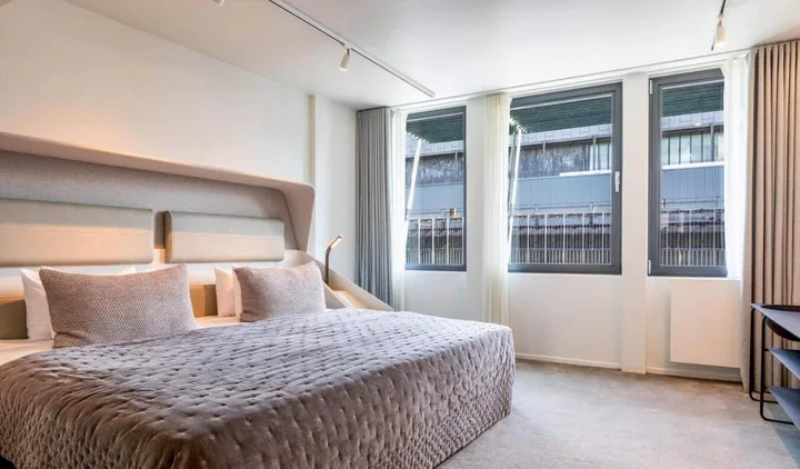 Contemporary hotel bedroom with large upholstered bed, track lighting, and floor-to-ceiling windows.