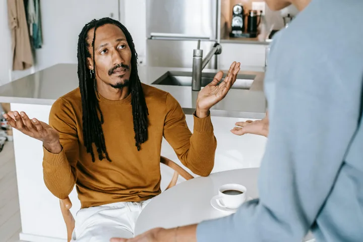 Man with dreadlocks gesturing with raised hands in a tense conversation at a kitchen counter.