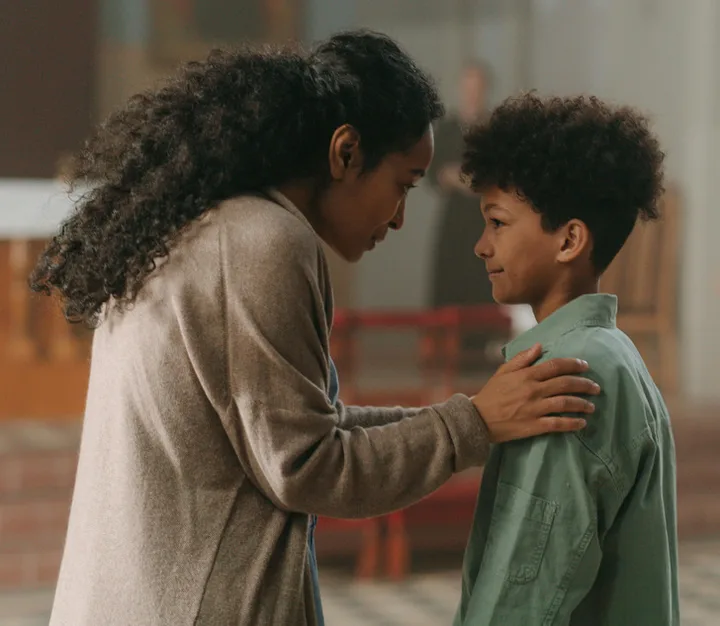 Woman tenderly placing hand on a young boy's shoulder in a warm, caring indoor moment.