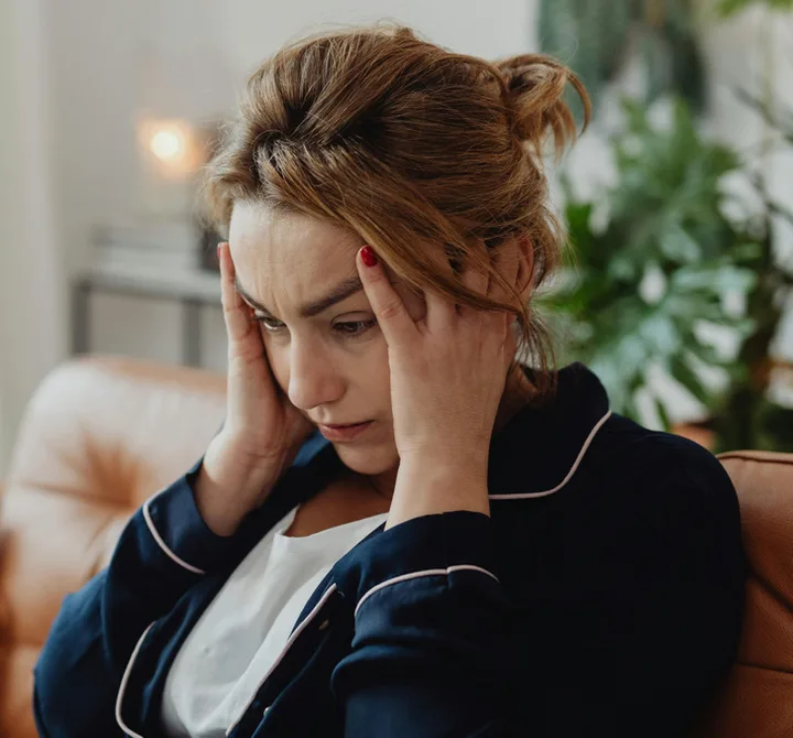 Woman sitting on a couch holding her head in her hands, looking stressed and distressed.