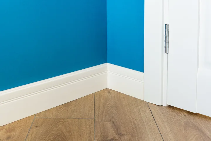 White baseboard molding in corner of blue-walled room with wood floor and white door hinge.