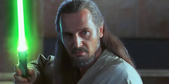 Qui-Gon Jinn holding an ignited green lightsaber, looking alert in a warmly lit interior.