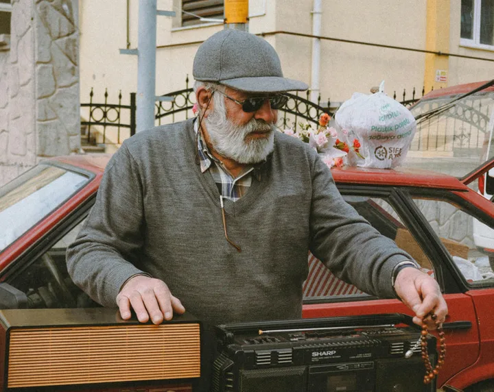 Older bearded man in a cap and sunglasses handling vintage electronics at what appears to be an outdoor market.