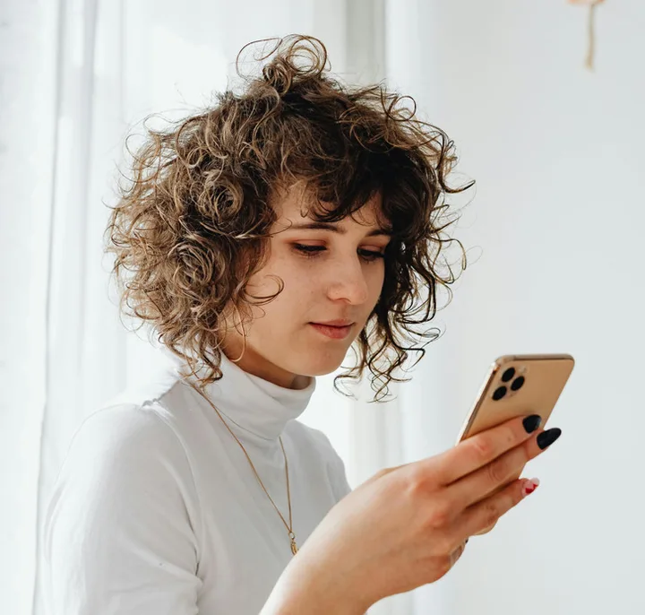 Young woman with curly hair looking down at her smartphone with a neutral expression.