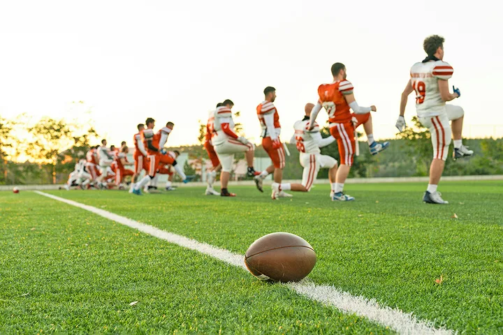 Football players in orange and white uniforms warming up on a green field at sunset.