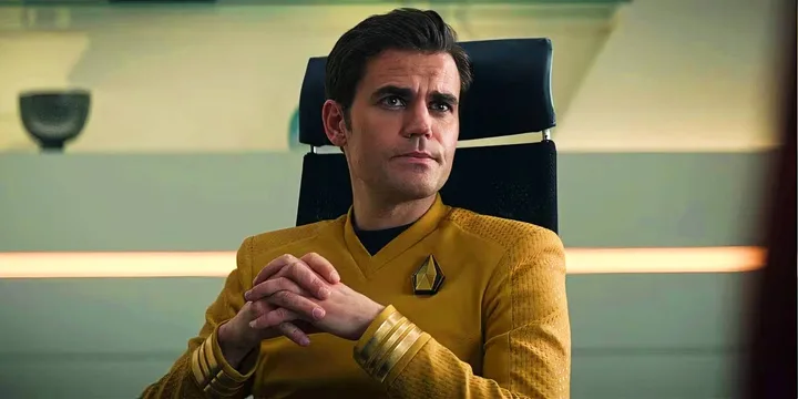 Male actor in gold Star Trek uniform seated in command chair with hands clasped.