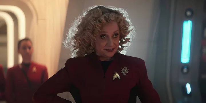 Female actor in red Star Trek uniform smiling in a futuristic corridor.