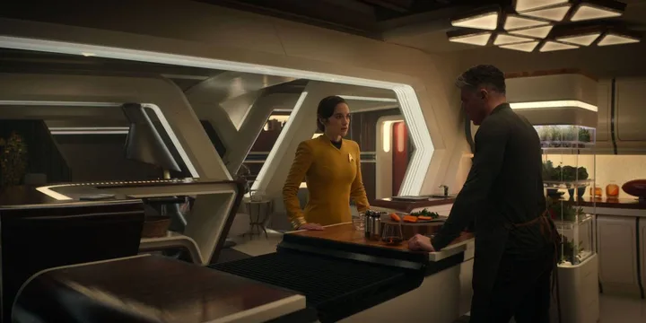 Female officer in gold Star Trek uniform talks to a man in a futuristic kitchen setting.