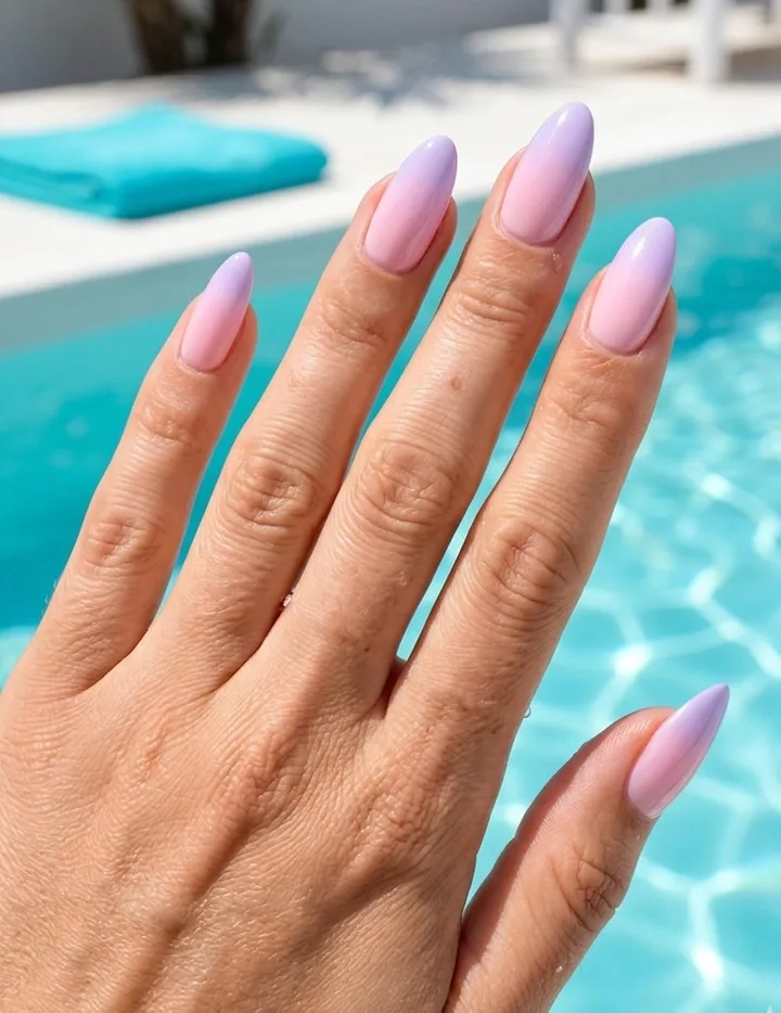 Close-up of almond-shaped nails with pastel purple-pink ombre finish, held near a blue swimming pool.