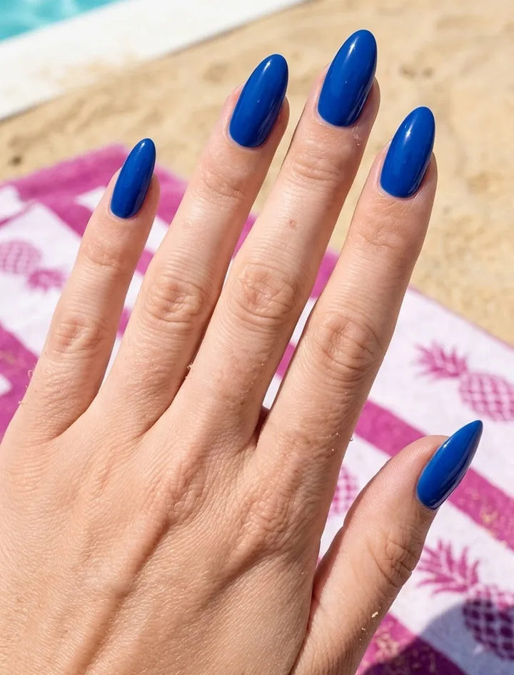 Hand with long almond nails painted bold cobalt blue, resting on a pineapple-print beach towel.