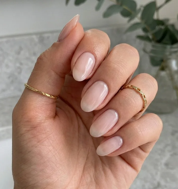 Hand with sheer nude glossy almond nails wearing two delicate gold rings, soft neutral background.