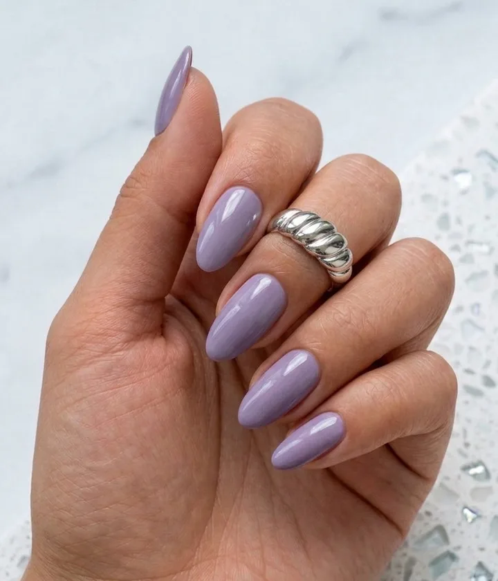 Hand with glossy lavender almond nails wearing a chunky silver ring on a marble surface.