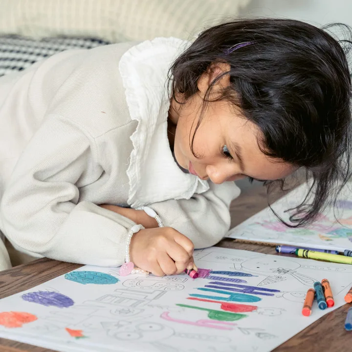 Young girl lying on a table intensely coloring a drawing with crayons, focused and engaged.