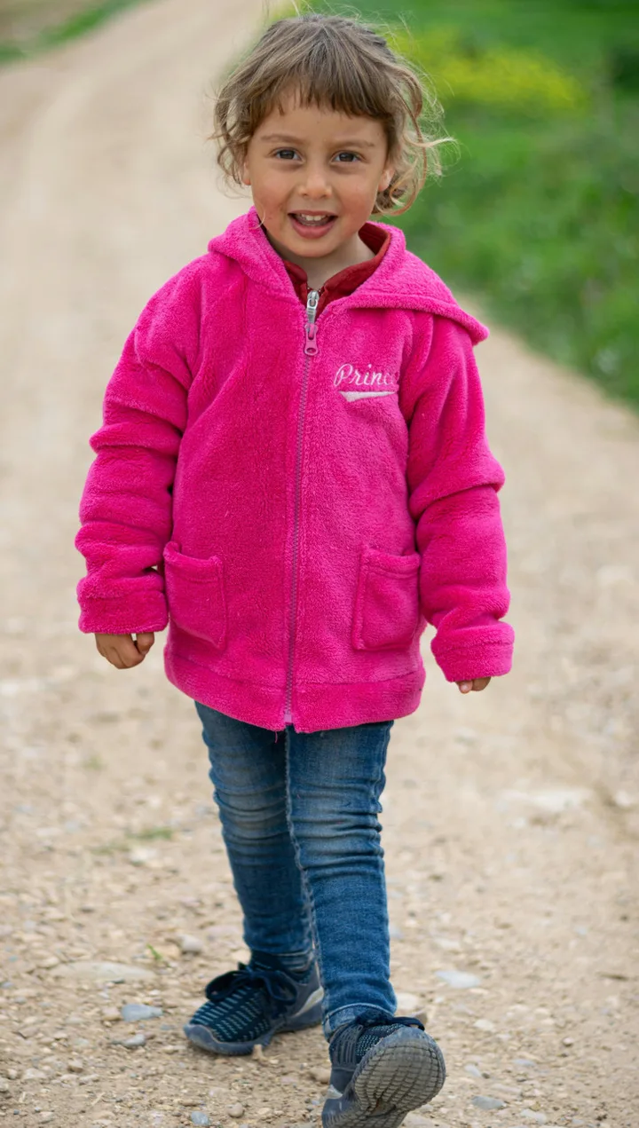 Cheerful toddler girl in bright pink fleece jacket walking along a dirt path outdoors.