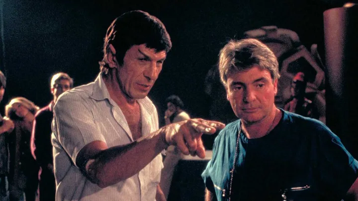 Two men on a dark film set, one with pointed ears pointing direction to the other.