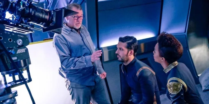 Director with glasses giving instructions to two actors in Star Trek uniforms on a sci-fi set.