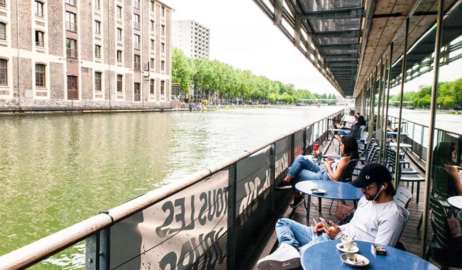 People relaxing at an outdoor cafe terrace alongside a canal with industrial brick buildings opposite.