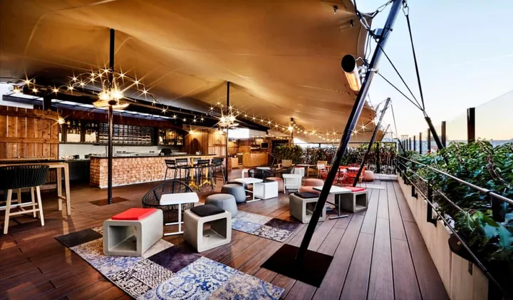 Covered rooftop bar terrace with string lights, lounge seating, and a city skyline view at dusk.