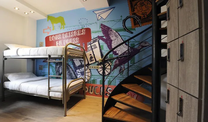 Colorful hostel dorm with Paris-themed mural, bunk bed, spiral staircase, and storage lockers.