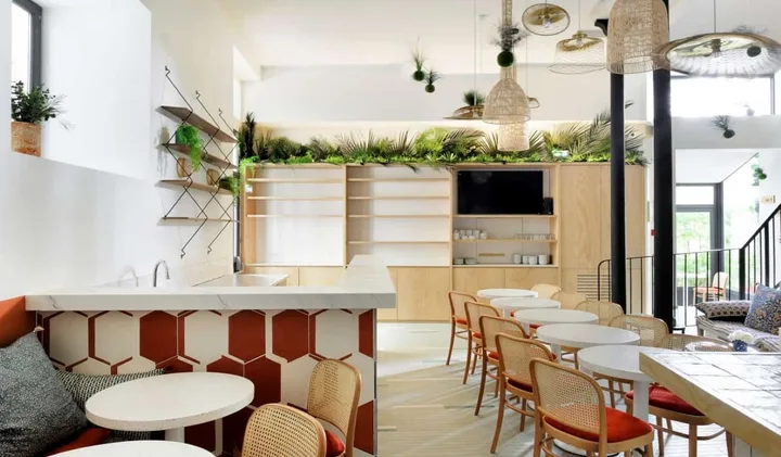 Bright modern hostel common area with rattan chairs, plants, hexagonal tiles, and kitchen counter.