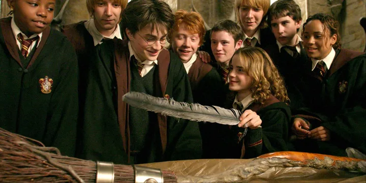 Group of Hogwarts students in robes gathered around a broomstick, smiling in a classroom scene.